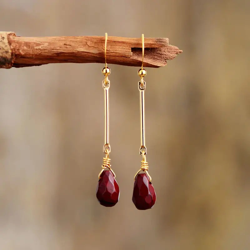 Gold earrings with red teardrop stones hanging on a branch against a blurred natural background