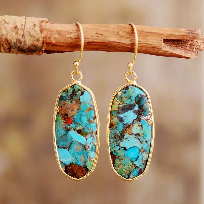 Turquoise earrings with gold frames hanging on a wooden stick against a blurred natural background