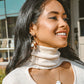Woman wearing a beige turtleneck sweater with hoop earrings and a necklace, smiling outdoors.