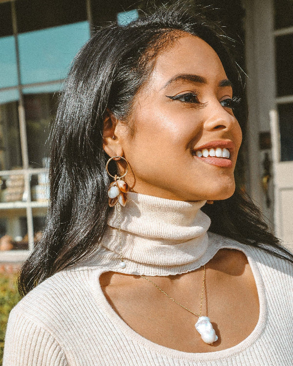 Woman wearing a beige turtleneck sweater with hoop earrings and a necklace, smiling outdoors.