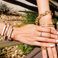 Close-up of a hand wearing multiple gold bracelets and rings with a blurred natural background