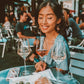 Woman sitting at a table with wine glasses and food, smiling outdoors.