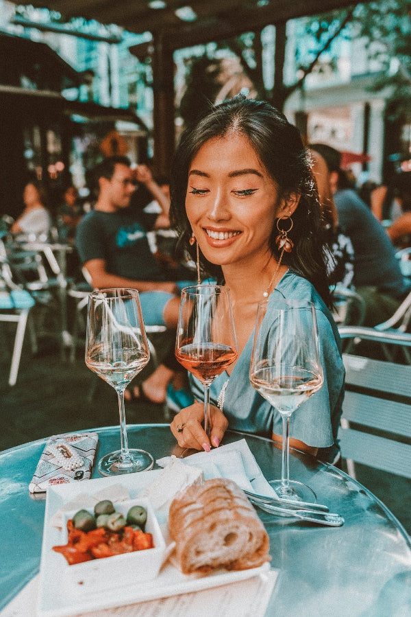 Woman sitting at a table with wine glasses and food, smiling outdoors.