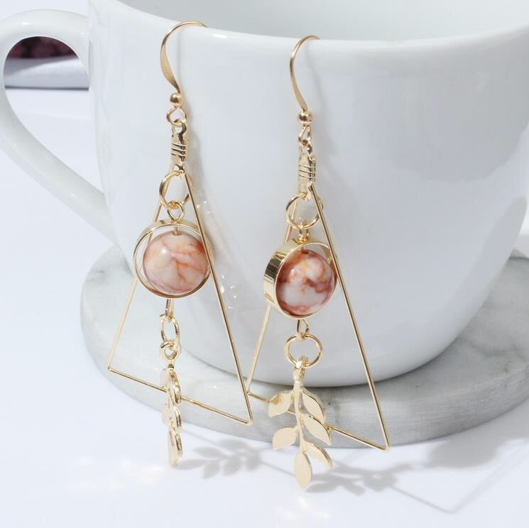 Gold earrings with pink stones and leaf charms in front of a white mug.