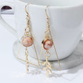 Gold earrings with pink stones and leaf charms in front of a white mug.