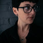 Person wearing glasses and a necklace against a dark brick wall