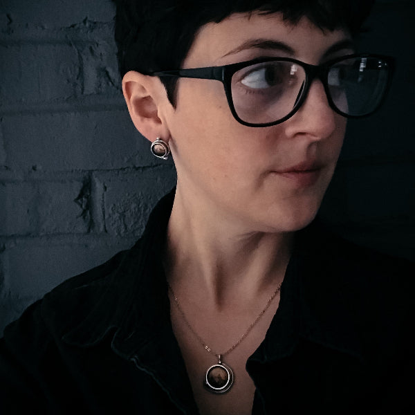 Person wearing glasses and a necklace against a dark brick wall