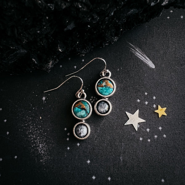 Pair of earrings with turquoise and moonstone stones on a dark surface with star decorations.