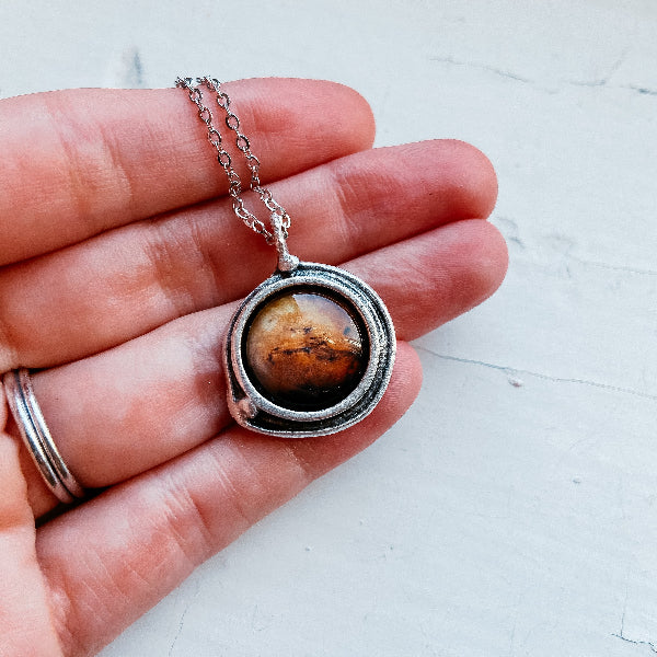 Hand holding a necklace with a pendant featuring a mountain landscape.