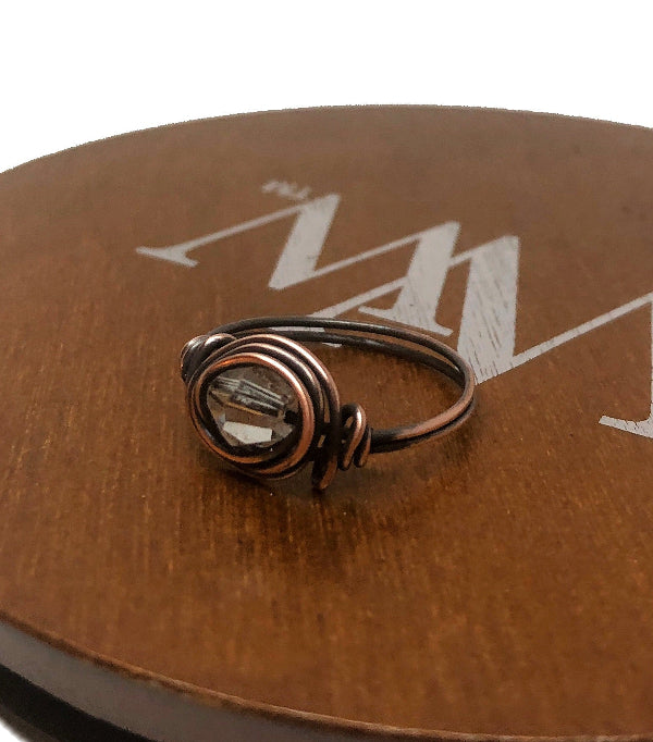 Bronze ring with a clear gemstone on a brown leather surface with a brand logo.