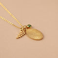 Gold necklace with fingerprint charm, angel wing charm, and green gemstone on a beige background