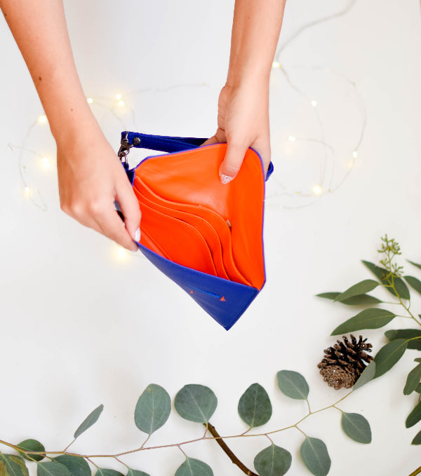 Orange and blue clutch held by a person with a white background and green leaves.