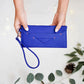 Blue clutch bag held by a person with a white background and green leaves.