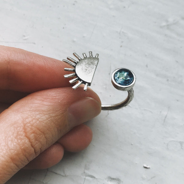 Silver ring with a sun design and gemstone held by a hand on a light gray background