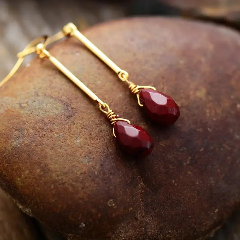 Gold earrings with red teardrop beads on a textured stone surface