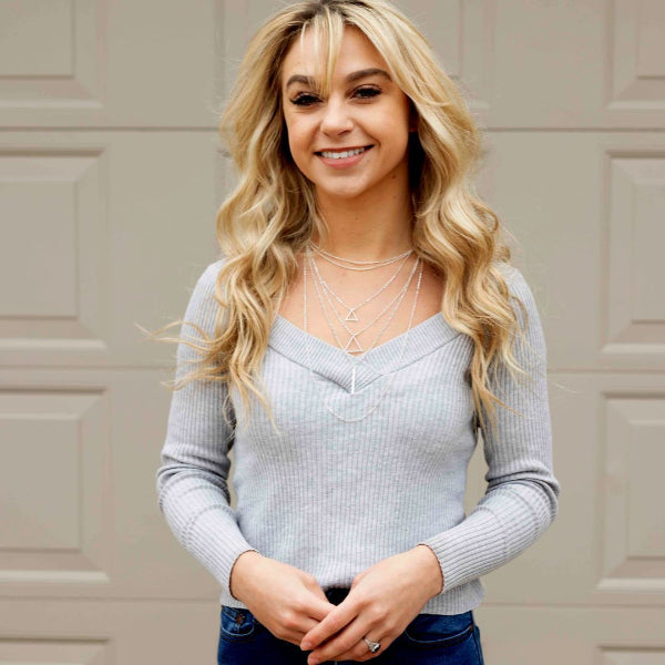 Woman wearing a light gray sweater with layered necklaces in front of a beige wall.