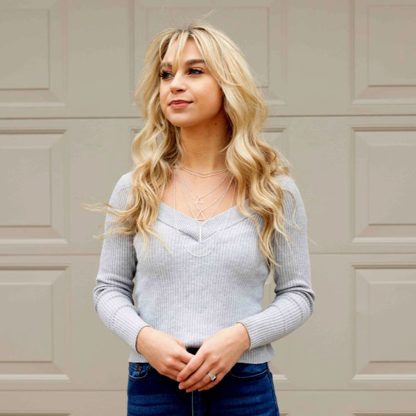 Woman with blonde hair wearing a light gray sweater and blue jeans against a beige wall.
