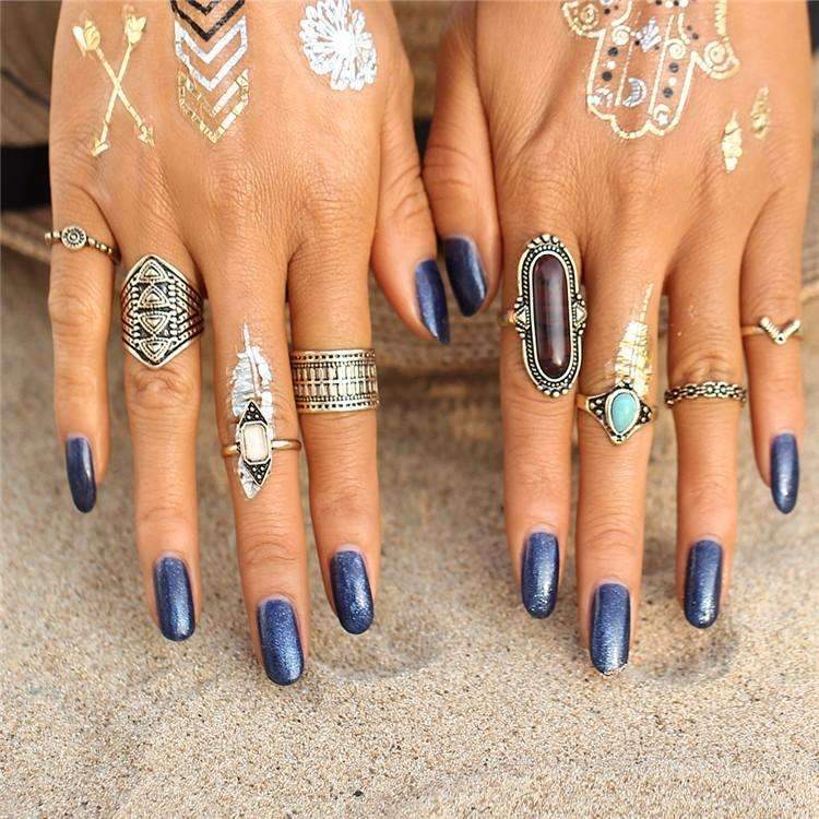 Close-up of hands with multiple rings and blue nail polish on a sandy background