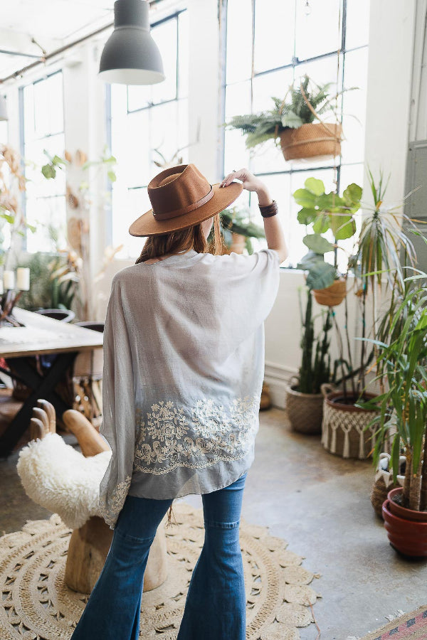 Person wearing a white lace shawl and brown hat in a room with plants and a rug.