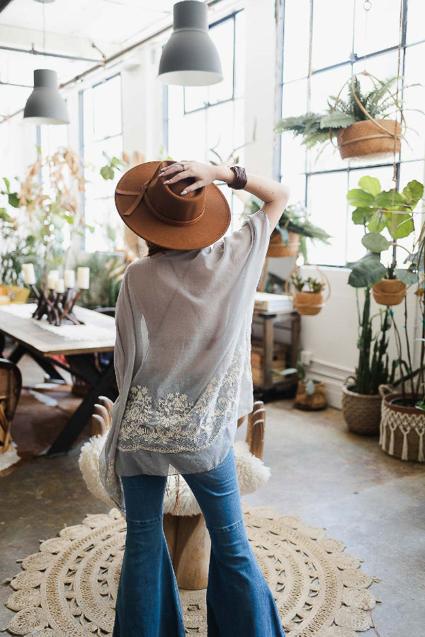 Person wearing a brown hat and white top with lace details in a plant-filled room.